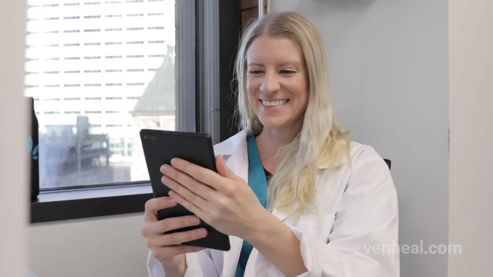 A clinician in a white coat smiles at a tablet during a telehealth visit, a frame from the Veriheal commercial