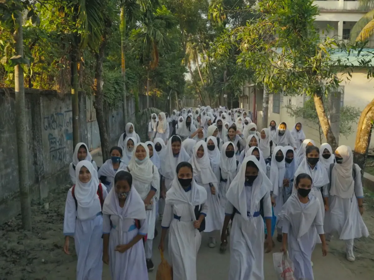 Twenty schoolgirls in white uniforms walking together down a tree-lined road toward camera, from the She Creates Change episode Keya