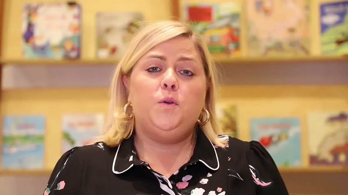 A woman speaking to camera in front of a shelf of children’s picture books, from a Room to Read policy video